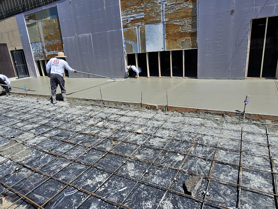 Finisher floating a new concrete slab beside a building with rebar set for the next pour, concrete work