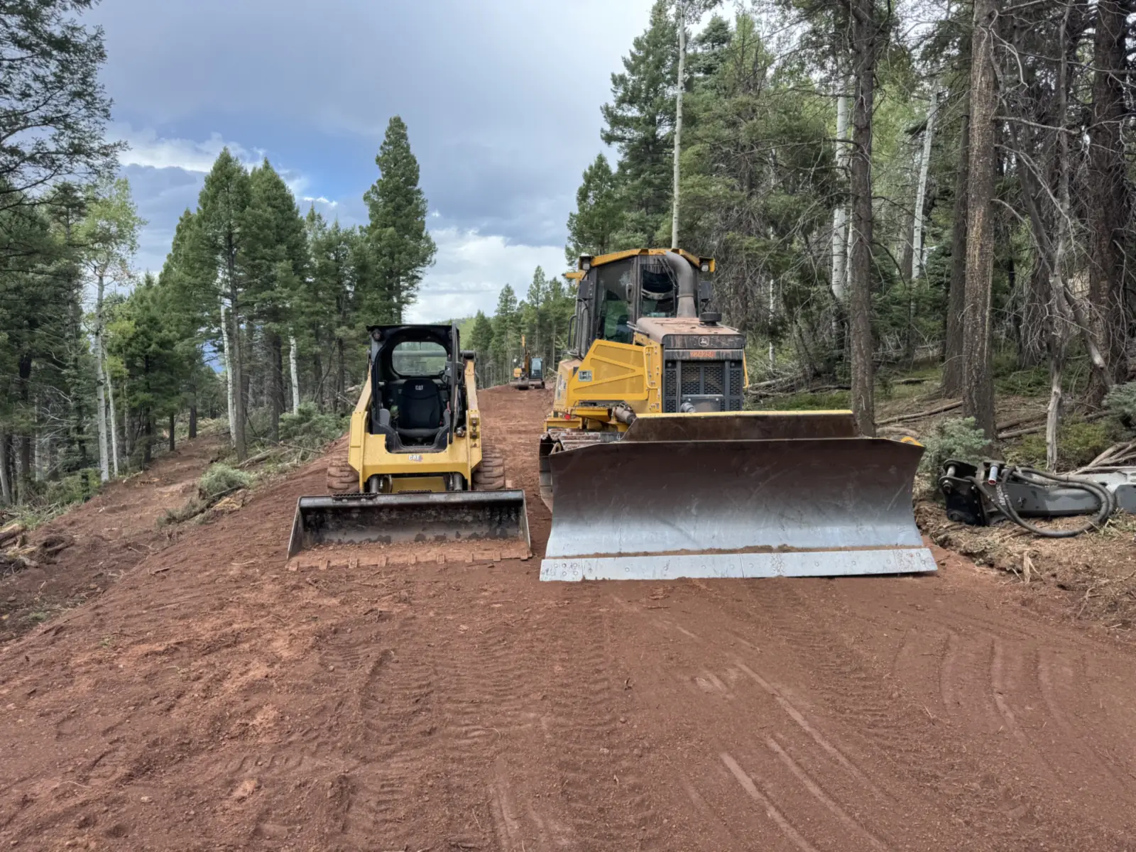 Compact track loader and dozer parked on a freshly cut lane through pines with an excavator beyond, land clearing
