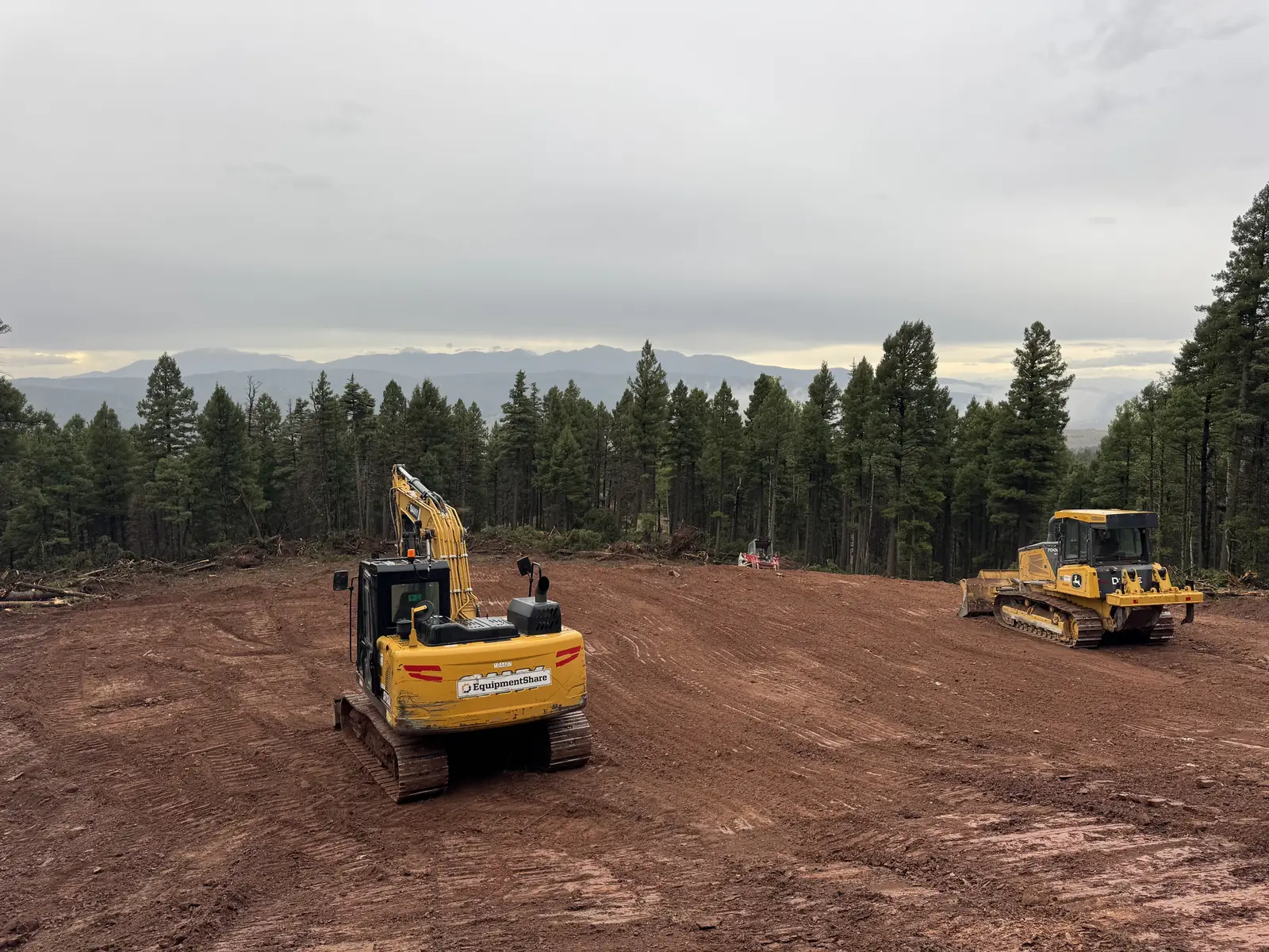 Excavator and dozer on freshly cleared ground ringed by pines with mountains behind, land clearing