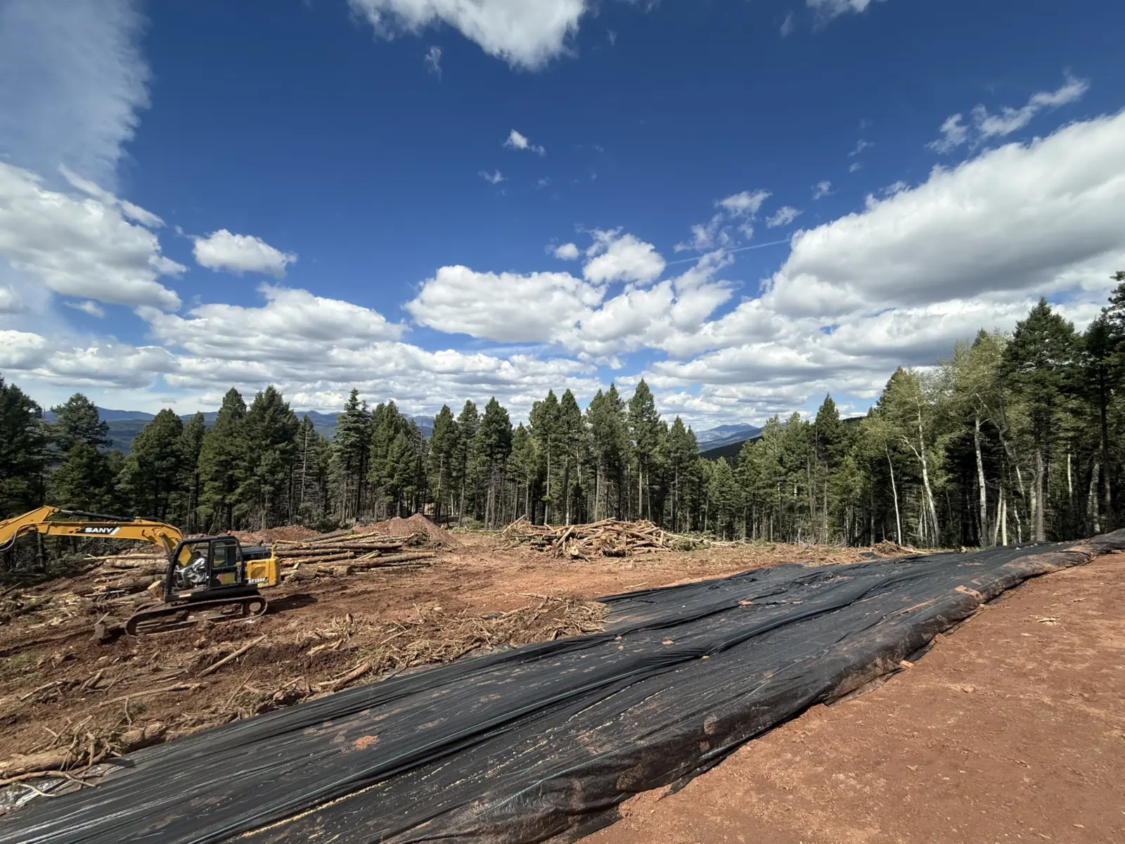 Excavator beside decked logs and brush piles with erosion sheeting along the cleared edge, land clearing