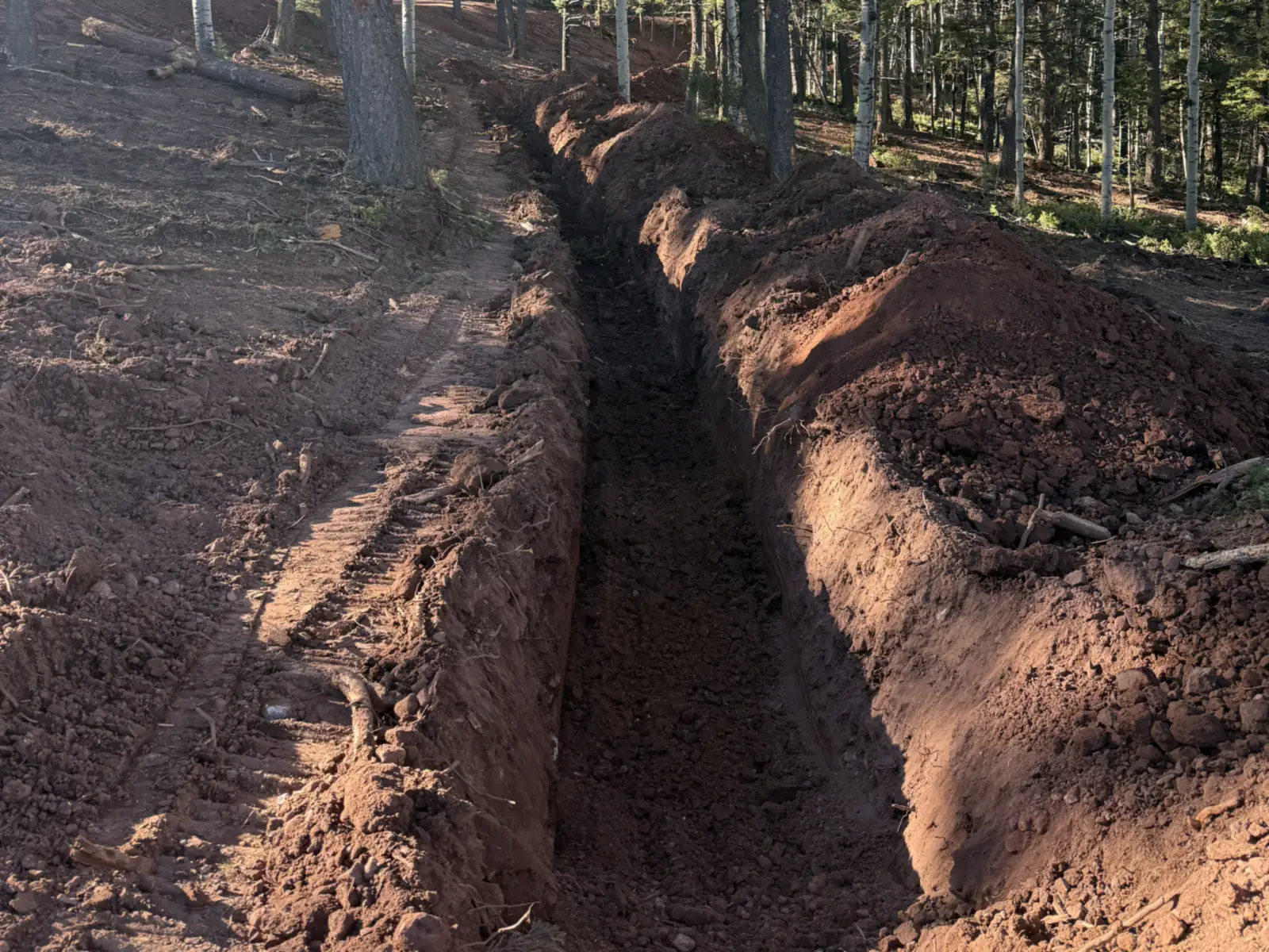 Narrow open trench dug through red soil with the spoil piled along one side, utility trenching