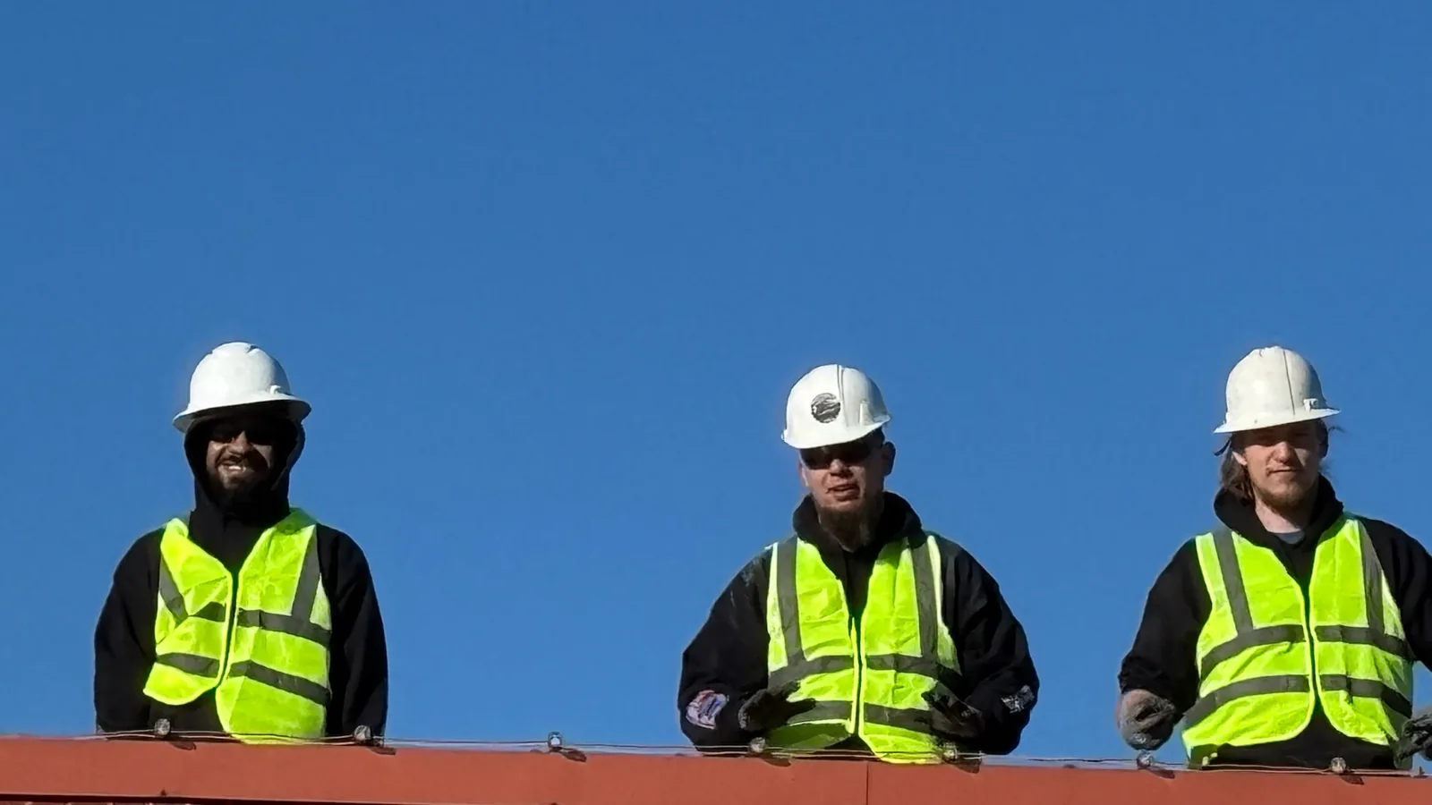 Three crew members in hard hats and safety vests on a roof edge